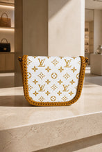 Load image into Gallery viewer, A Louis Vuitton shoulder bag with a white monogram canvas body and a mustard yellow braided leather trim, displayed on a light beige stone ledge in a luxury boutique setting.