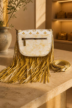 Load image into Gallery viewer, A cream and gold crossbody bag with a long fringe skirt and a matching coiled strap, displayed on a stone counter in a warm, sunlit boutique setting.