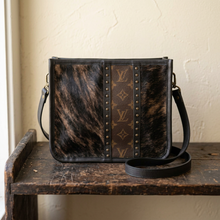 Load image into Gallery viewer, A crossbody bag with a brown and black brindle cowhide pattern, a central brown monogram canvas stripe, and brass studs. Cowhide is unique and may vary slightly.