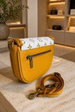 Load image into Gallery viewer, A mustard yellow leather shoulder bag with a white flap featuring a gold monogram pattern, displayed on a light stone surface with its matching strap in front.

