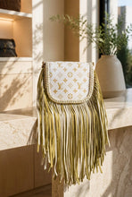 Load image into Gallery viewer, A Louis Vuitton shoulder bag with a white leather flap covered in gold monogram print, featuring a braided gold trim and long cascading gold fringe, displayed on a marble counter in a warm, sunlit interior.
